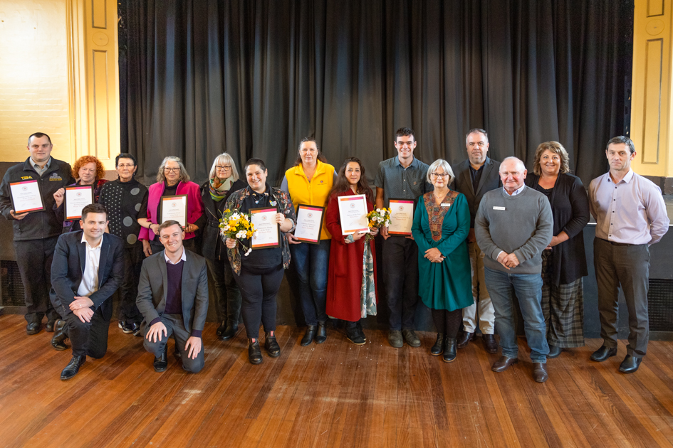 A group of people standing indoors posing for a photo. Some individuals hold framed certificates, while one holds a bouquet of flowers. They are standing in front of a black curtain on a wooden floor. Everyone is smiling.