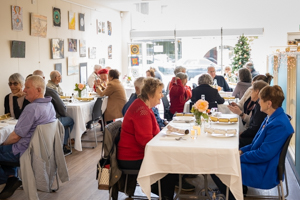 A group of people dining in a cozy restaurant. The tables are set with white tablecloths and flowers. Paintings decorate the light-colored walls, and a Christmas tree is visible near the window. The atmosphere is lively and social.