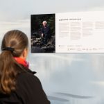 A person with long hair and a scarf reads an informational display titled The Black Swan featuring a photograph of someone kneeling by rocks. The background is a light, softly blurred pattern.