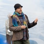 A person wearing a plaid shirt, checkered blazer, colorful scarf, and black beanie speaks outdoors. They appear animated, gesturing with one hand. The background features a person in a gray jacket and a blue, cloud-like backdrop.