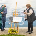 A man unveils a plaque on an easel beside a waterfront mural while a woman holding papers speaks into a microphone. Yellow flowers rest at the base. The background depicts a water scene with gentle waves.