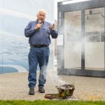 An older man in a blue shirt and jeans speaks into a microphone outdoors. He stands near a small smoking pot with greenery, next to a black container labeled ARTBOX. A large mural with abstract blue patterns is in the background.