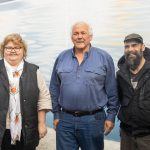 Three people standing in front of a mural with a water-themed background. The first person is wearing a black vest and scarf, the second a blue shirt and jeans, and the third a dark jacket and cap. All are smiling at the camera.