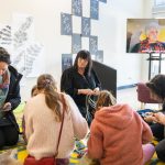 A group of people is gathered around a colorful rug, engaging in an activity inside a room. Artworks are displayed on the walls, and a screen shows a person speaking. Everyone appears focused on the task at hand.
