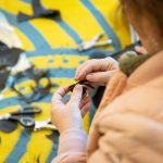 A person meticulously crafts a small leather object by hand, surrounded by scattered crafting materials on a vibrant blue and yellow surface. Various tools like scissors and scrap pieces are visible, indicating detailed handiwork.