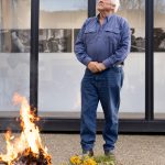 An older man with white hair stands on a patio, looking up thoughtfully. He is wearing a blue button-up shirt and jeans. In front of him, a small fire burns in a bowl, surrounded by yellow flowers and greenery.