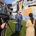A person speaks at a press event outdoors, with a mural of black swans and an orange sun in the background. Two reporters, one holding a camera and the other taking notes, capture the scene on a grassy area.