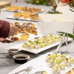 A person in a red jacket holds a plate, selecting food from a buffet. The table features sushi rolls, tartlets, and various appetizers. Small dishes of soy sauce accompany the sushi. A floral arrangement decorates the setting.