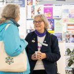 Two women are talking in front of a wall covered with colorful flyers. One wears a blue coat and holds a tote bag, while the other wears glasses and a navy jacket, holding a cup. A potted plant is visible nearby.