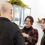 A group of four people are standing indoors, engaged in conversation. A woman with dark hair in a plaid coat and another with lighter hair in a brown jacket are smiling. A man with a bald head is seen from the back. A display case is in the background.
