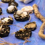 Small black pouches filled with seashells and stones are arranged on a blue patterned cloth. A piece of driftwood lies to the right, and strands of natural fibers accent the display.