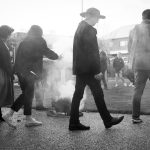 A group of people walking outdoors in a line, surrounding a smoking fire pit. The scene is captured in black and white, with a mix of men and women dressed in casual and formal attire. Some figures are partially obscured by smoke.