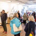 A diverse group of people socializes and observes artwork in a gallery. The space is bright with artworks displayed on walls. People engage in conversation and some children are present, creating a lively atmosphere.