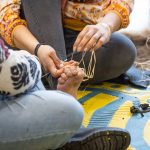 Two people sitting cross-legged on a colorful mat, working together to weave or tie something around one persons bare foot. Various tools are scattered on the mat. They are focused on their task, which involves string or cords.