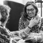 Two women sitting and smiling in conversation. One wears glasses, a scarf, and a floral top, while the other wears a patterned sweater. They appear to be knitting in a cozy setting. Black and white image.
