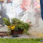 A person in jeans stands next to a smoking container filled with green pine branches and yellow flowers on the ground. Nearby, children in colorful outfits are partially visible, one holding a piece of paper.