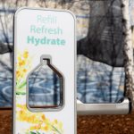 A water bottle refill station with the words Refill, Refresh, Hydrate is in focus. It features floral designs around a bottle-shaped opening. In the blurred background, theres a mural depicting marine life.