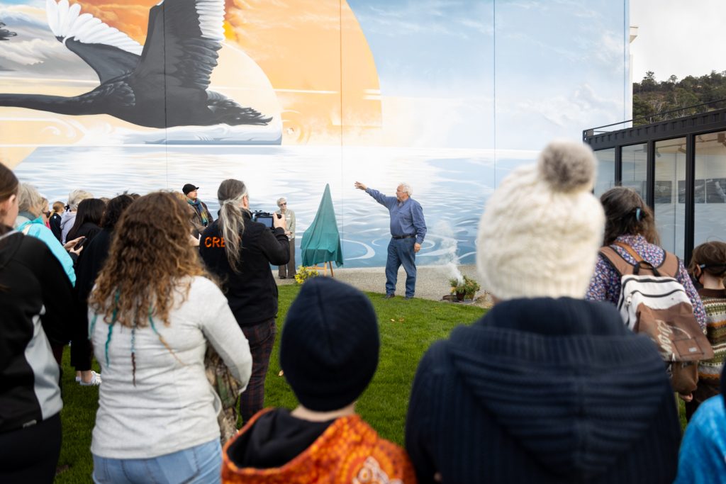 A group of people gather outdoors facing a speaker in front of a large mural featuring a bird and a sun. The mural is painted on a building wall. The event appears casual, with attendees wearing jackets and hats.