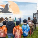 A group of people stand outside in front of a large mural depicting an eagle flying over water with a sun in the background. Some people wear colorful jackets. They are gathered on a grassy area near a modern building with large windows.
