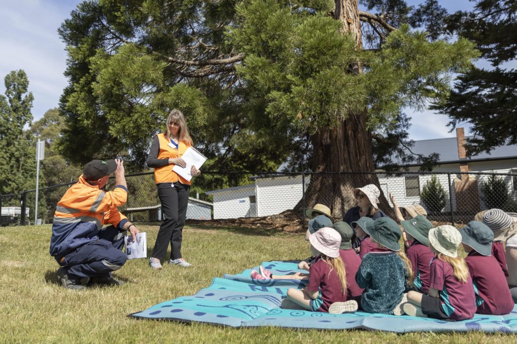 A woman and a man in orange safety vests are speaking to a group of children seated on mats outside. The children are wearing hats and school uniforms. Theyre gathered under a large tree on a grassy area.