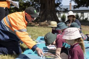 A man in an orange jacket hands a booklet to a child wearing a maroon uniform and hat. Other children in similar uniforms are sitting on a blue mat outdoors. A woman in the background wears a sunhat and sunglasses.
