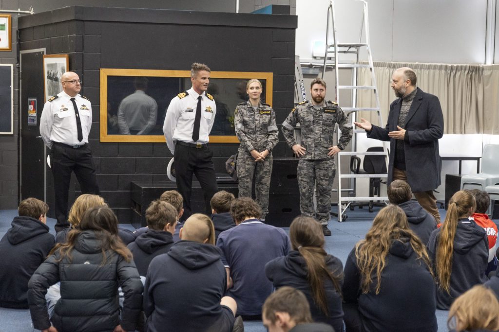 A group of students sit on the floor facing four military personnel and a man in a suit. The military personnel wear uniforms, two in white and two in camouflage. They are inside a room with black walls and a ladder.