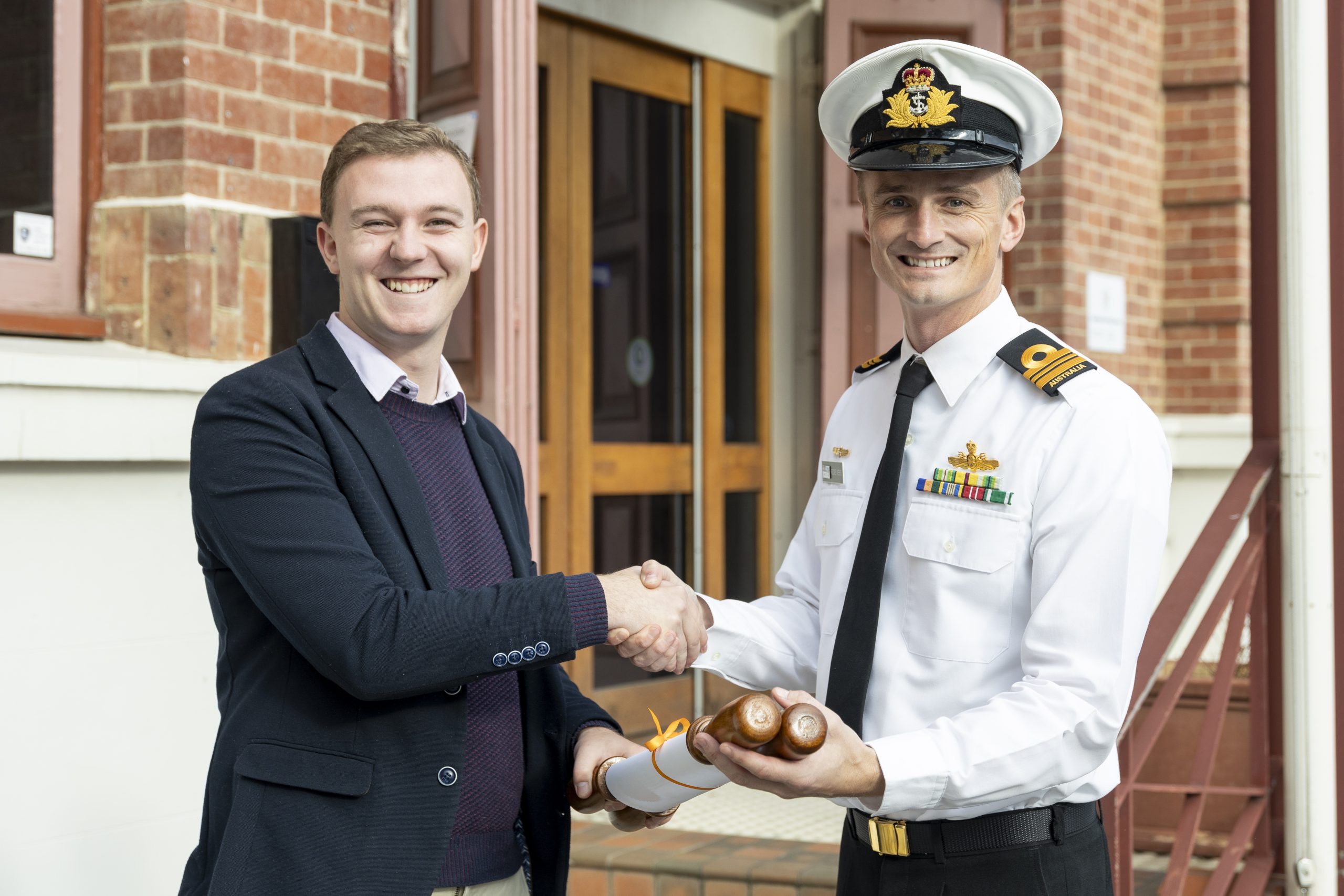 A smiling man in civilian clothing shakes hands with a uniformed naval officer holding a wooden object wrapped with ribbon. They are standing outside a brick building with wooden doors.