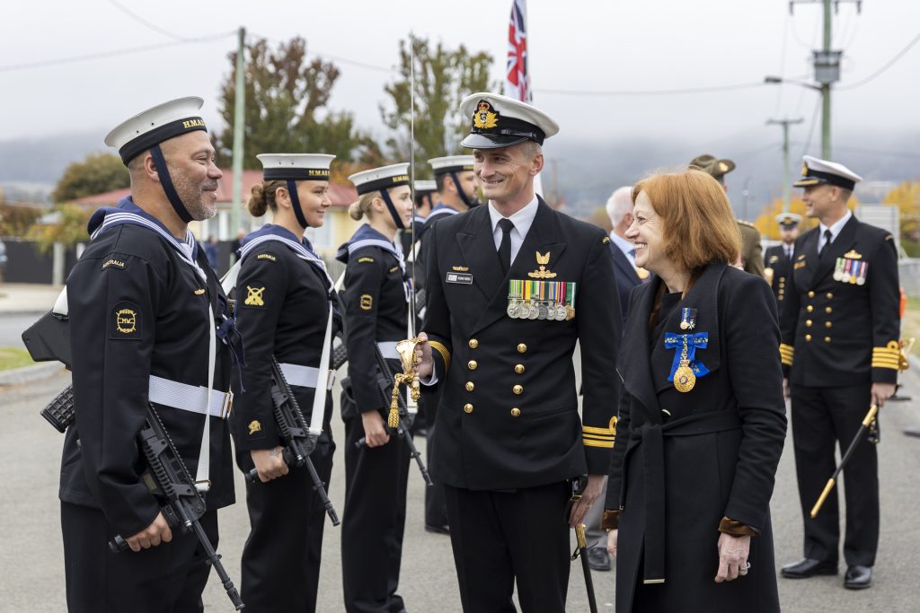 A naval officer in uniform, with medals and a sword, converses with a woman wearing a black coat and a blue cross medal. Other uniformed personnel stand in formation in the background, holding rifles. Flags are visible in the overcast setting.