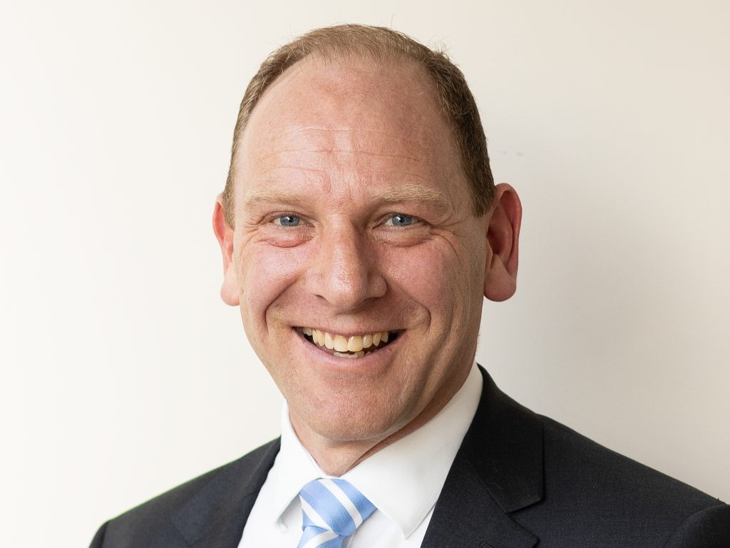 A man in a suit and tie smiling at the camera against a plain white background.
