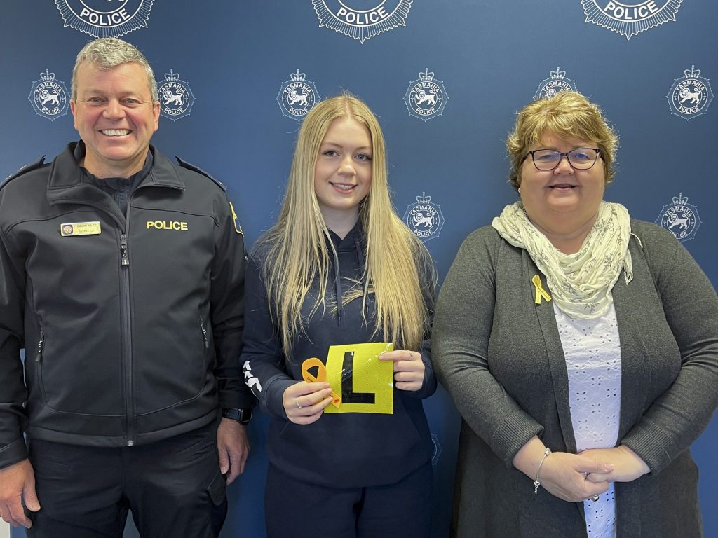 Three people stand in front of a police-themed backdrop. The person in the center holds a yellow learners permit symbol. All are smiling and wearing clothing with badges or symbols.