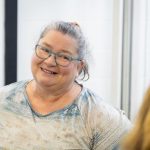A woman with glasses and a patterned shirt smiles warmly while looking at someone off-camera. She has gray hair pulled back and stands in a bright room with a window in the background.