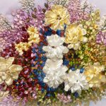 A vibrant embroidery of a flower bouquet with cream, white, and yellow satin roses. Surrounding are pink, red, blue, and green leaves and small flowers on a light fabric. The textures create a three-dimensional effect.
