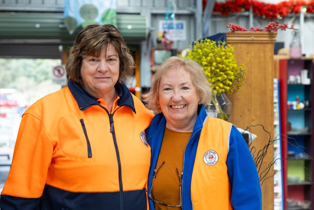 Two women wearing bright orange and blue work jackets smile at the camera inside a colorful, cluttered shop or workshop. They appear to be employees or volunteers, equipped with name tags and emblems on their jackets.