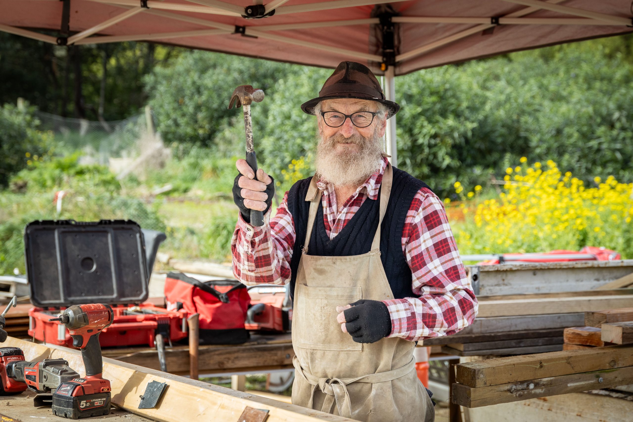 Elderly man with a beard, wearing a hat, glasses, and plaid shirt, smiles while holding a hammer under a tent canopy. Surrounded by tools, he stands at a wooden workbench with a lush green garden in the background.