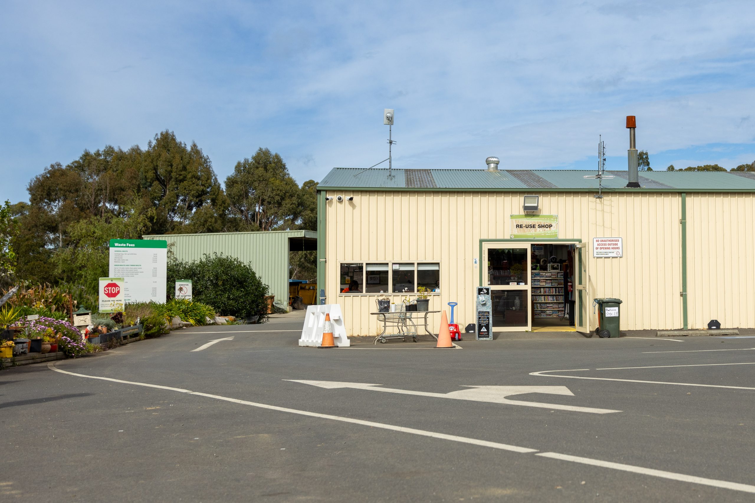 A garden center with a beige industrial building featuring a large open door. Outside, there are traffic cones, tables, and potted plants. A signboard is visible on the left, and a clear blue sky is overhead.