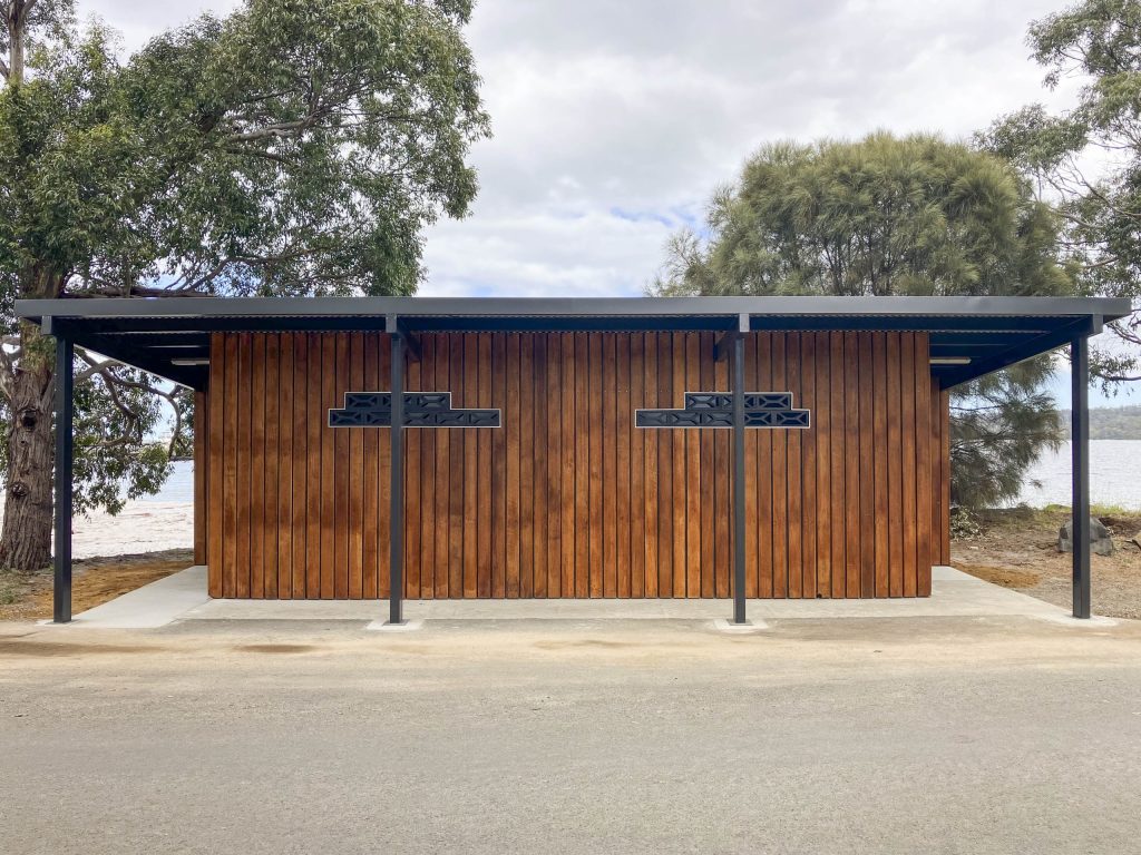 A modern public restroom facility with a simple design featuring vertical wooden paneling and a flat roof. It is situated outdoors with trees on either side and a view of a lake in the background.