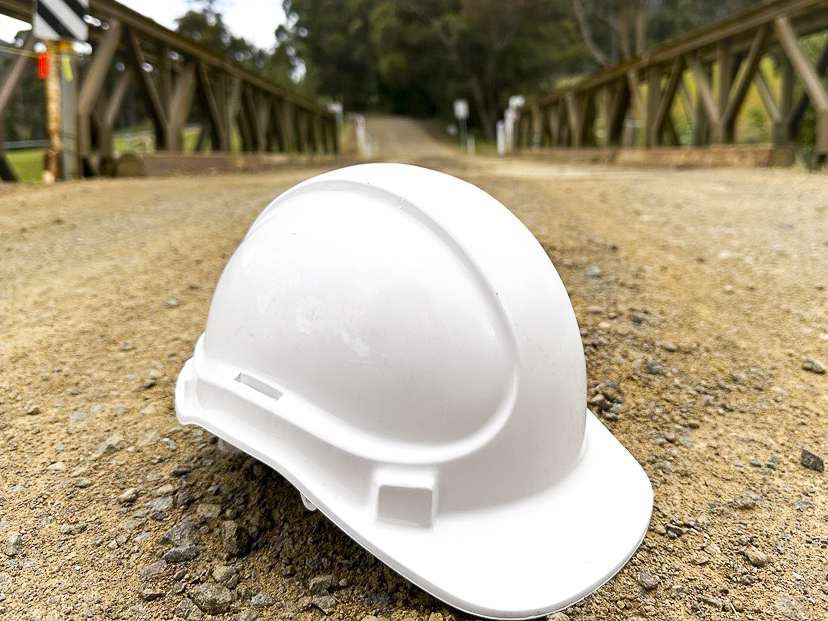 A white hard hat lies on a dirt pathway, leading to a wooden bridge. The bridge is framed by lush greenery under an overcast sky, creating a serene and slightly rustic atmosphere.
