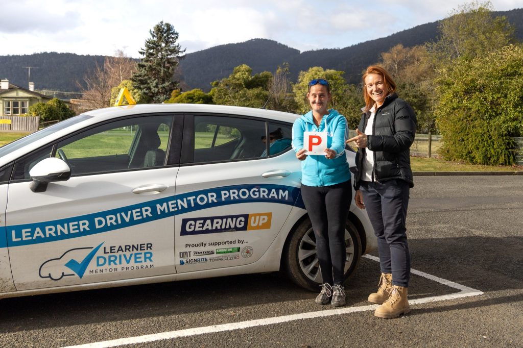 Two people standing next to a white car with Learner Driver Mentor Program branding. One is holding a red P plate. They are smiling, with mountains and trees in the background.