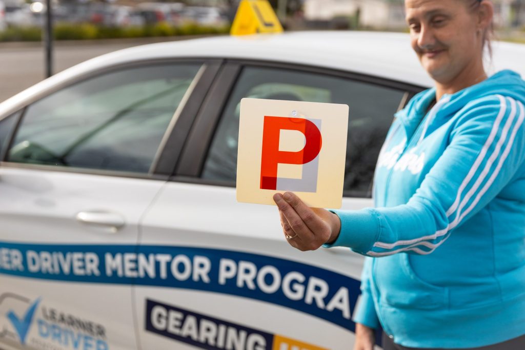 A person in a blue hoodie holds a red P plate in front of a white car labeled with Learner Driver Mentor Program. The car has a yellow sign on top. The setting appears to be an outdoor parking area.