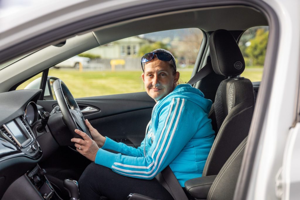 A person wearing a blue jacket sits in the drivers seat of a car, looking at the camera. The car door is open, and theres a grassy field visible in the background.