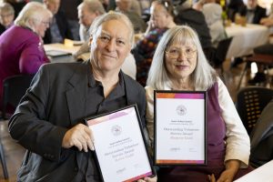 A man and woman sit close together at a crowded event. They are each holding framed certificates that read Outstanding Volunteer Service Award. Both are smiling at the camera. The background shows people seated at tables.