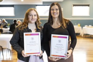 Two people stand indoors holding framed certificates. Both are smiling. The person on the left has long hair and wears a black top. The person on the right has long hair and wears a dark top. Tables and chairs are visible in the background.