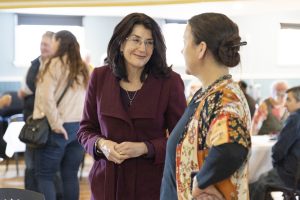 Two women are conversing in a room filled with people. One wears a burgundy coat, the other a colorful patterned outfit with a lanyard. Others in the background are blurred, sitting and standing around tables.