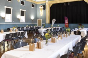 A decorated banquet hall with long tables covered in white cloths, small brown gift bags, flowers, and papers. Rows of black chairs surround the tables. Banners are near a stage with a black curtain, and the room has light blue walls and tall windows.