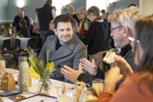A man in a gray coat is engaged in conversation with two others at a table in a crowded indoor setting. The table has a small plant, a water bottle, and food boxes. People are mingling in the background.