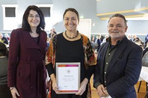 Three people stand together indoors, smiling. The person in the center is holding a framed Citizen of the Year certificate. A group of people is visible in the background seated at tables.