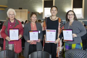Four women stand side by side, each holding a framed certificate. They are smiling and dressed in colorful attire, standing in a room with blue walls and black chairs.