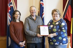 Three people stand smiling indoors in front of several flags. The person in the center holds an award plaque. The two beside them stand closely and smile. The background features red curtains and white walls.