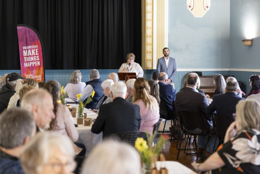 A person speaks at a podium in a crowded room with tables. Attendees listen attentively. A banner reads Volunteers Make Things Happen. A man stands nearby. Tulip centerpieces decorate the tables. The room has a high ceiling and elegant decor.