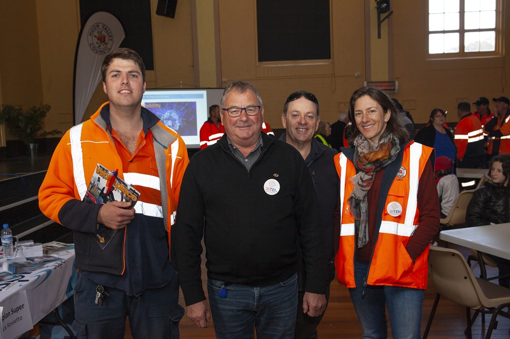 Four people stand together indoors wearing safety vests and casual attire. Theyre smiling, and behind them is a group of people in matching outfits. There are tables with brochures and other materials nearby.